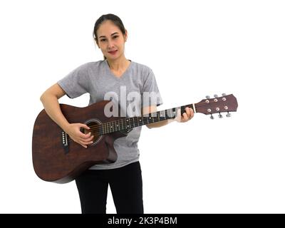 Asiatische Frau in grauem T-Shirt und schwarzer Hose, die akustische Gitarre spielt. Porträt auf weißem Hintergrund mit Studiolicht. Stockfoto