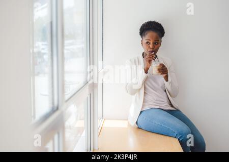 Afrikanische Frau im Café sitzt auf der Fensterbank und trinkt Eiskaffee mit einem Stroh. Lächelnde junge Geschäftsfrau. Genießen Sie den Morgen im Café. Stockfoto