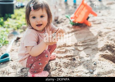 Liebenswert kleines Mädchen auf Spielplatz im Sandkasten entdecken neue Dinge in der Natur. Sensorische Entwicklung für Kinder im Freien. Stockfoto