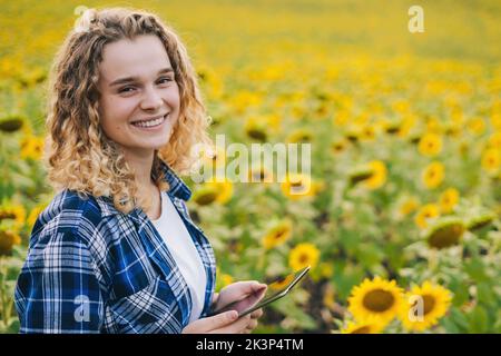 Ein Farmerporträt blickt auf die Kamera, hält ein Tablet in den Händen und prüft ihr Sonnenblumenfeld. Intelligente Landwirtschaft. Landwirtschaftliche Technologien. Ländlich Stockfoto
