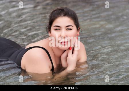 Übergroße junge Brünette Erwachsenen-Modell in schwarzen einteiligen Badeanzug liegend und Entspannung in Mineral-Thermalwasser im Außenpool im Spa Stockfoto