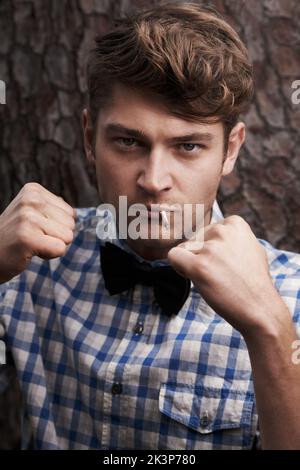 Style mit einer rauen Kante. Porträt eines schlauen jungen Mannes mit erhobenen Armen in einer kämpfenden Pose, der vor einem Baum steht. Stockfoto