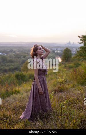 Schöne junge lächelnde Mädchen in einem langen braunen Kleid steht entlang des Rasens. Glückliche Frau geht bei Sonnenuntergang auf einem Hügel mit Blick auf den Fluss. Konzept des havi Stockfoto