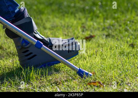 Junge mit gebrochenem Fuß und orthopädischem Schuh oder Spaziergänger nach Knochenbruch ruht im öffentlichen Park auf einer Bank im grünen Gras, um neu zu erschaffen und zu rehabilitieren Stockfoto