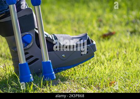 Junge mit gebrochenem Fuß und orthopädischem Schuh oder Spaziergänger nach Knochenbruch ruht im öffentlichen Park auf einer Bank im grünen Gras, um neu zu erschaffen und zu rehabilitieren Stockfoto