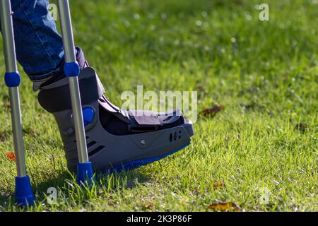 Junge mit gebrochenem Fuß und orthopädischem Schuh oder Spaziergänger nach Knochenbruch ruht im öffentlichen Park auf einer Bank im grünen Gras, um neu zu erschaffen und zu rehabilitieren Stockfoto