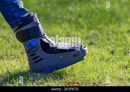 Junge mit gebrochenem Fuß und orthopädischem Schuh oder Spaziergänger nach Knochenbruch ruht im öffentlichen Park auf einer Bank im grünen Gras, um neu zu erschaffen und zu rehabilitieren Stockfoto