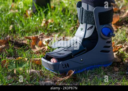 Junge mit gebrochenem Fuß und orthopädischem Schuh oder Spaziergänger nach Knochenbruch ruht im öffentlichen Park auf einer Bank im grünen Gras, um neu zu erschaffen und zu rehabilitieren Stockfoto