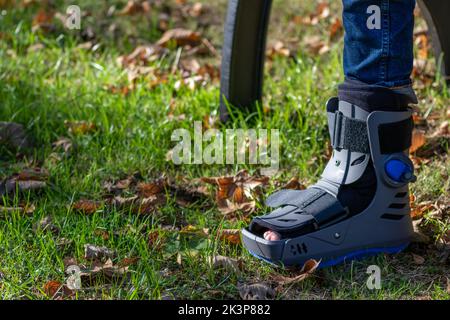 Junge mit gebrochenem Fuß und orthopädischem Schuh oder Spaziergänger nach Knochenbruch ruht im öffentlichen Park auf einer Bank im grünen Gras, um neu zu erschaffen und zu rehabilitieren Stockfoto