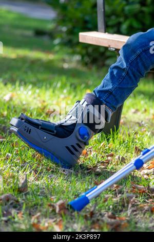 Junge mit gebrochenem Fuß und orthopädischem Schuh oder Spaziergänger nach Knochenbruch ruht im öffentlichen Park auf einer Bank im grünen Gras, um neu zu erschaffen und zu rehabilitieren Stockfoto