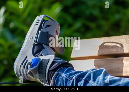 Junge mit gebrochenem Fuß und orthopädischem Schuh oder Spaziergänger nach Knochenbruch ruht im öffentlichen Park auf einer Bank im grünen Gras, um neu zu erschaffen und zu rehabilitieren Stockfoto