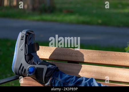 Junge mit gebrochenem Fuß und orthopädischem Schuh oder Spaziergänger nach Knochenbruch ruht im öffentlichen Park auf einer Bank im grünen Gras, um neu zu erschaffen und zu rehabilitieren Stockfoto