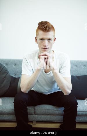 Ernsthafte Kontemplation. Ein rothaariger Mann, der auf einer Couch sitzt und direkt auf die Kamera blickt. Stockfoto