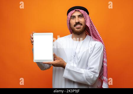 Arabischer Geschäftsmann in traditioneller Kleidung mit leerem digitalen Tablet vor orangefarbenem Hintergrund Stockfoto