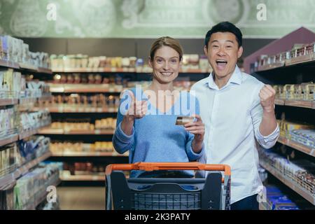 Familieneinkäufe. Junge schöne interracial paar Einkaufen in einem Supermarkt. Glückliche asiatische Frau und Mann stehen mit Wagen, zeigen Kreditkarte, zufrieden mit Einkaufen, Preise, Rabatte. Stockfoto