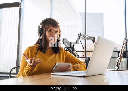 Schöne weibliche Podcasterin in Kopfhörern und Brillen gestikulieren während der Aufnahme Audio-Podcast-Studio, interviewen einen Gest, Blick auf.Junge hübsche Frau in Kopfhörern im Gespräch in ein Mikrofon Stockfoto