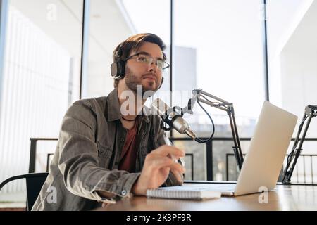 Junger Mann Host in Kopfhörer und Brillen sitzen am Tisch, Streaming Audio-Podcast mit Mikrofon und Laptop im Studio, halten einen Stift, in Richtung. Social Media, Podcasting, Blogging-Konzept Stockfoto