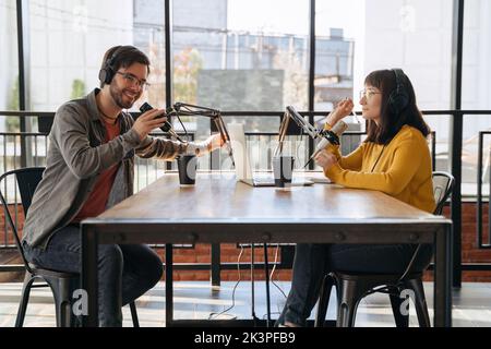 Zwei fröhliche Gastgeber lächeln, während sie in der Nähe von Mikrofonen sitzen und im Sendestudio sprechen und Podcast aufnehmen. Junge Frau interviewt einen Mann für Audio-Podcast auf ihrem Broadcast-Kanal Stockfoto