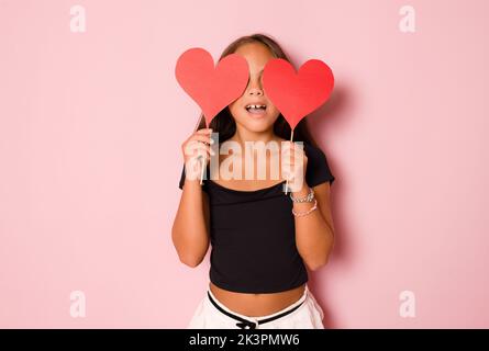 Foto von charmanten süßen jungen Mädchen tragen elegante Party-Kleidung halten roten Papier Herzen isoliert rosa Farbe Hintergrund Stockfoto