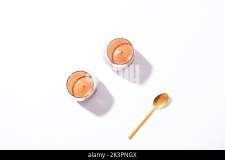 Zwei Gläser mit Schokoladenmousse und einem Löffel im Sonnenlicht auf weißem Hintergrund. Frühstück am Morgen, veganes Essen. Draufsicht, flach liegend. Stockfoto