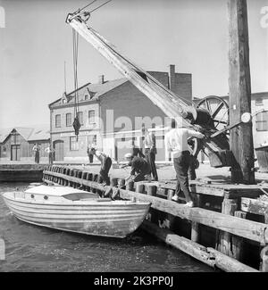 1950s Boot. Der Start eines hölzernen Segelbootes. Der alt aussehende Kran hat das Boot im Wasser abgesenkt und sein Haken hält das Segelboot nicht mehr. Dieses Modell war das beliebte Volksboot. Der Besitzer muss nun den Mast und das Rig montieren. Solvesborg Schweden 1954 Kristoffersson Ref. BX31-2 Stockfoto