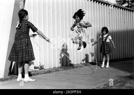 Mexiko im Jahr 1960s. Drei Mädchen spielen auf der Straße, mit einem Springseil - einem Springseil. Ein Mädchen an jedem Ende des Seils schwingt es und ein Mädchen in der Mitte springt darüber. Mexiko 1964. Roland Palm Ref.-Ordner 2. Stockfoto