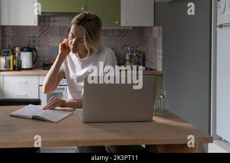 Beschäftigte Frauen mittleren Alters machen im Notebook Spuren und suchen über einen Laptop in der Küche nach bezahlter Arbeit. Stockfoto
