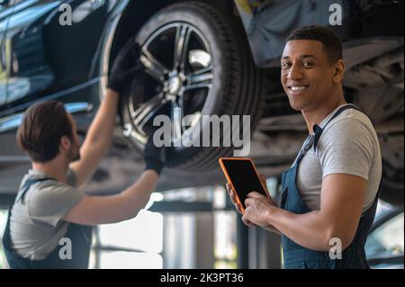 Lächelnder Automobiltechniker und sein Mitarbeiter reparieren das Fahrzeug Stockfoto