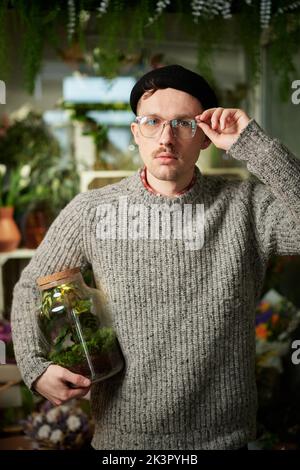 Attraktive bärtige männliche Holding-Glas mit Pflanze im Blumenlager. Gut aussehender Floristen-Mann mit Pullover, Mütze und Brille. Konzept für kleine Unternehmen. Hochwertiges vertikales Bild Stockfoto