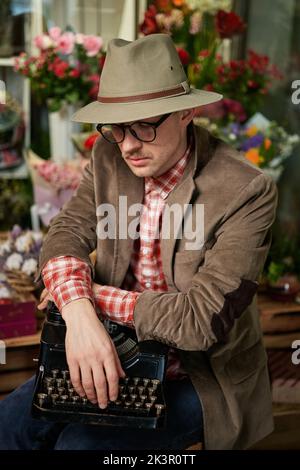 Attraktive kaukasische männliche Person in Brillen und Hut sitzend mit alter Schreibmaschine im Blumengeschäft denken. Dichter oder Schriftsteller Arbeitskonzept. Hochwertiges vertikales Bild Stockfoto