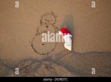 Roter Weihnachtsmann Hut auf einem weißen Stein und ein Schneemann auf Sand am Küstenstrand gemalt. Das Konzept der Feier von Weihnachtsferien während der Reise zu Stockfoto