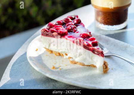 Eine einzelne Scheibe oder Portion frisch zubereiteter, nachgiebiger Kirschkäsekuchen. Es wird auf einem Teller mit einem Eiskaffee serviert. Zu einem Tisch im Freien in einem Café. Stockfoto