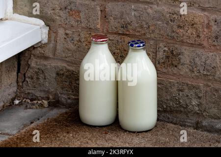 Zwei Pint Milch in Glasflaschen vor einer Haustür in Yorkshire, England. Die rote Oberseite ist fettarme Milch, die blaue Oberseite ist fetthalbleicht. Stockfoto