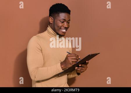 Portrait von attraktiven fokussierten trendy fröhlich Kerl Schreiben Liste Füllung Anwendung isoliert über braunen Hintergrund Stockfoto