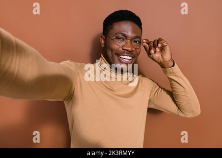 Selbstporträt von gutaussehenden trendy fröhlich Kerl Blogger posiert berührende Spezifikationen isoliert über braunen Hintergrund Stockfoto