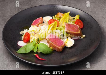 Leckerer Salat mit gebratenem Thunfisch, Gemüse und Wachtelei auf einem schwarzen Teller Stockfoto