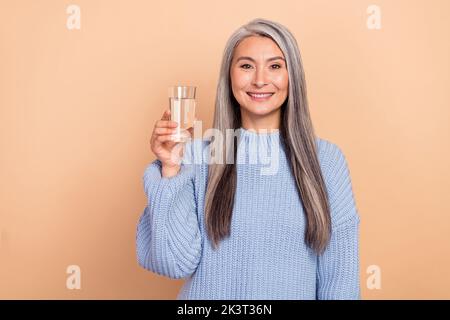 Foto von niedlichen liebenswert im Ruhestand Frau tragen blauen Pullover mit Glas Wasser isoliert beige Farbe Hintergrund Stockfoto