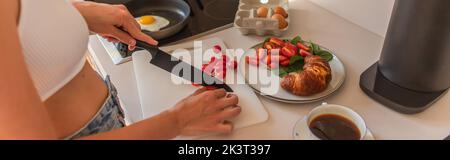 Beschnittene Ansicht einer Frau, die Kirschtomaten in der Nähe von Kaffee und Croissant in der Küche schneidet, Banner, Stockbild Stockfoto