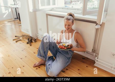 Junge Frau in Jeans hält Teller mit Ei und Croissant in der Nähe schottischer Falte Katze zu Hause, Stock Bild Stockfoto