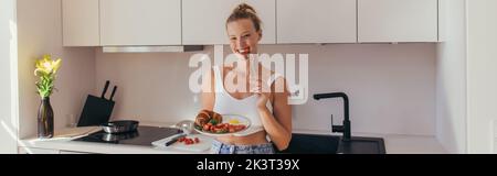 Positive blonde Frau, die Kirschtomate isst und Frühstück in der Küche hält, Banner, Stockbild Stockfoto