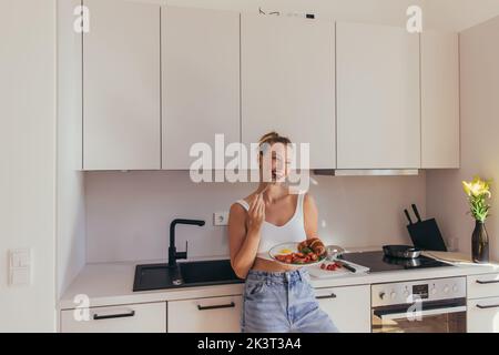 Lächelnde blonde Frau, die Kirschtomate isst und Teller mit Ei und Croissant in der Küche hält, Stockbild Stockfoto