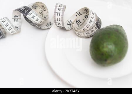 Kreative flache Lay-Komposition aus einer ganzen grünen Avocado und weichem Maßband auf einer weißen Platte isoliert auf Weiß. Stockfoto