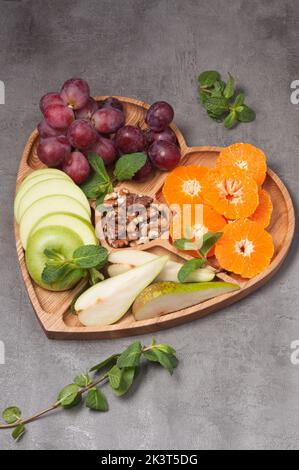 Herzförmiger Holzteller mit Früchten und Nussbaum auf grauem Hintergrund Stockfoto