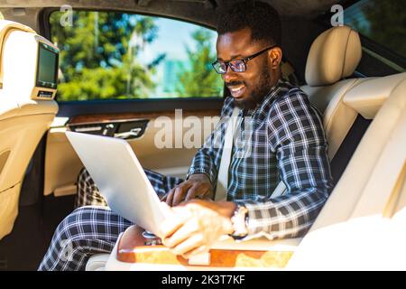 Gutaussehende erfolgreiche reiche afroamerikanische Geschäftsleute in einem stilvollen Anzug und sitzen in einem Luxusauto und arbeitet mit Laptop Stockfoto