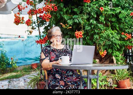 Positive ältere Dame in lässigem Outfit und Brille sitzt auf einem Stuhl am Tisch, während sie auf Netbook neben Tasse auf Untertasse und grünen Pflanzen mit Strömung surft Stockfoto