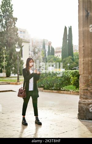 Ganzer Körper fokussierter hispanischer Frauen in stilvollem Outfit, der mit einer modernen Fotokamera in den Händen fotografiert, die mit grünen Pflanzen auf der Straße stehen Stockfoto
