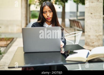 Seriöse hispanische Frau in eleganter Kleidung, die an einem modernen Netbook arbeitet, während sie auf der Terrasse des Cafés in der Stadt am Tisch sitzt Stockfoto