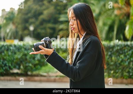 Fokussierte hispanische Frau in stilvollem Outfit, die mit einer modernen Fotokamera in den Händen fotografiert, die mit grünen Pflanzen auf der Straße stehen Stockfoto