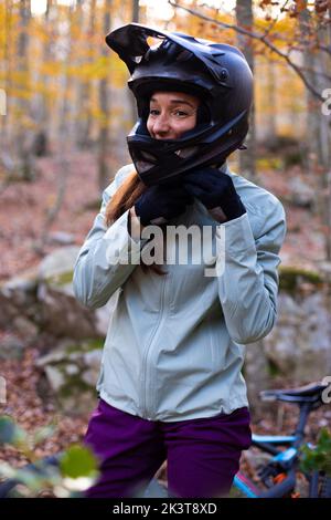 Glückliche Frau beim Anziehen und Befestigen von Schutzhelm, während sie in der Nähe des Fahrrads im Herbstwald steht Stockfoto