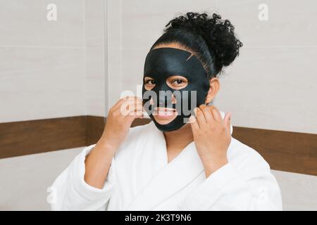 Charmante Frau in weißem Bademantel Anwendung feuchtigkeitsspendende schwarze Bettmaske während der Hautpflege Routine am Morgen Stockfoto